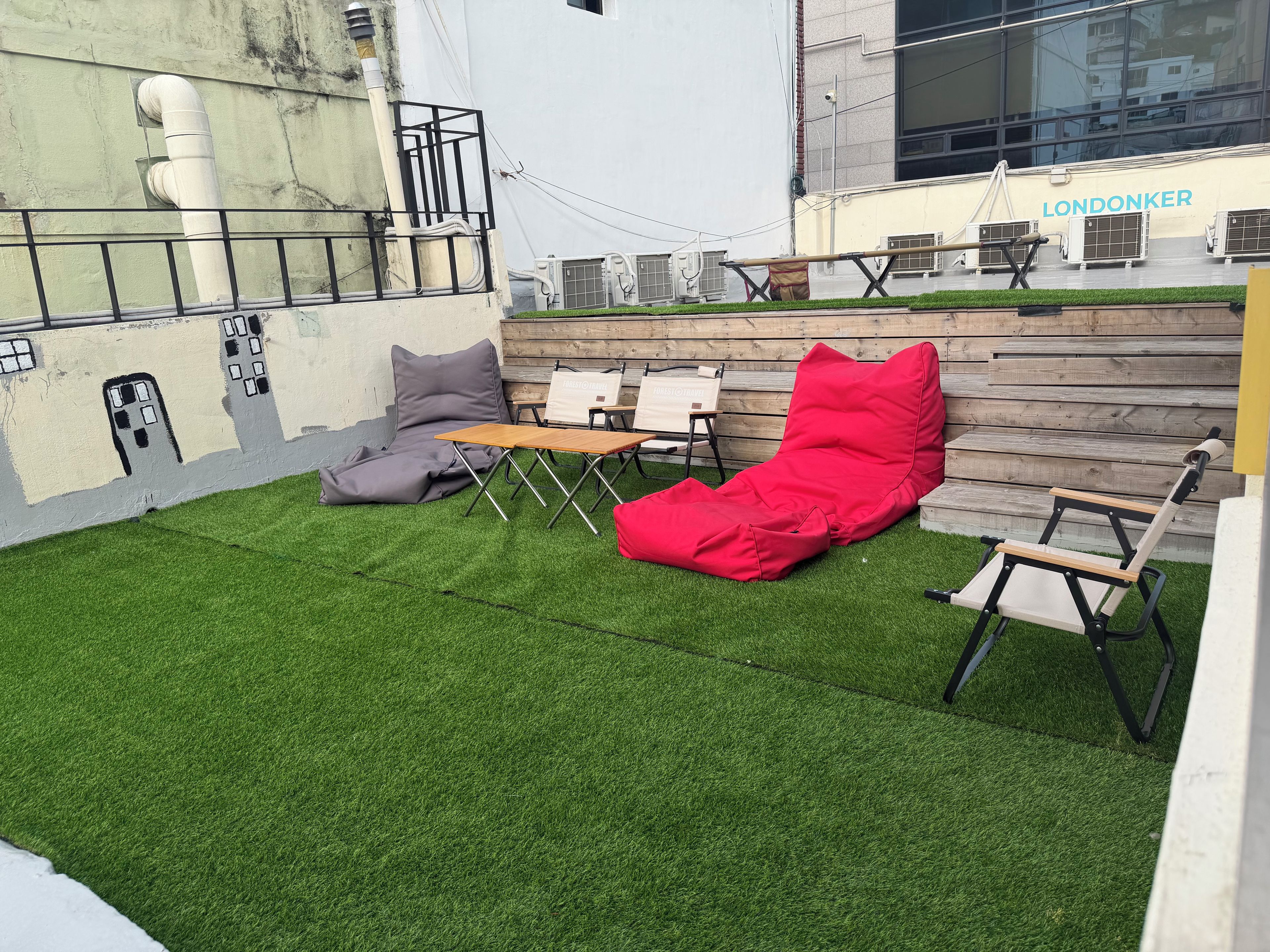 A rooftop terrace with artificial grass, a red lounge chair, a grey bean bag, and a wooden table.
