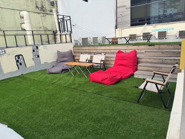A rooftop terrace with artificial grass, a red lounge chair, a grey bean bag, and a wooden table. - STAY zzZARM Hotel (Busan)