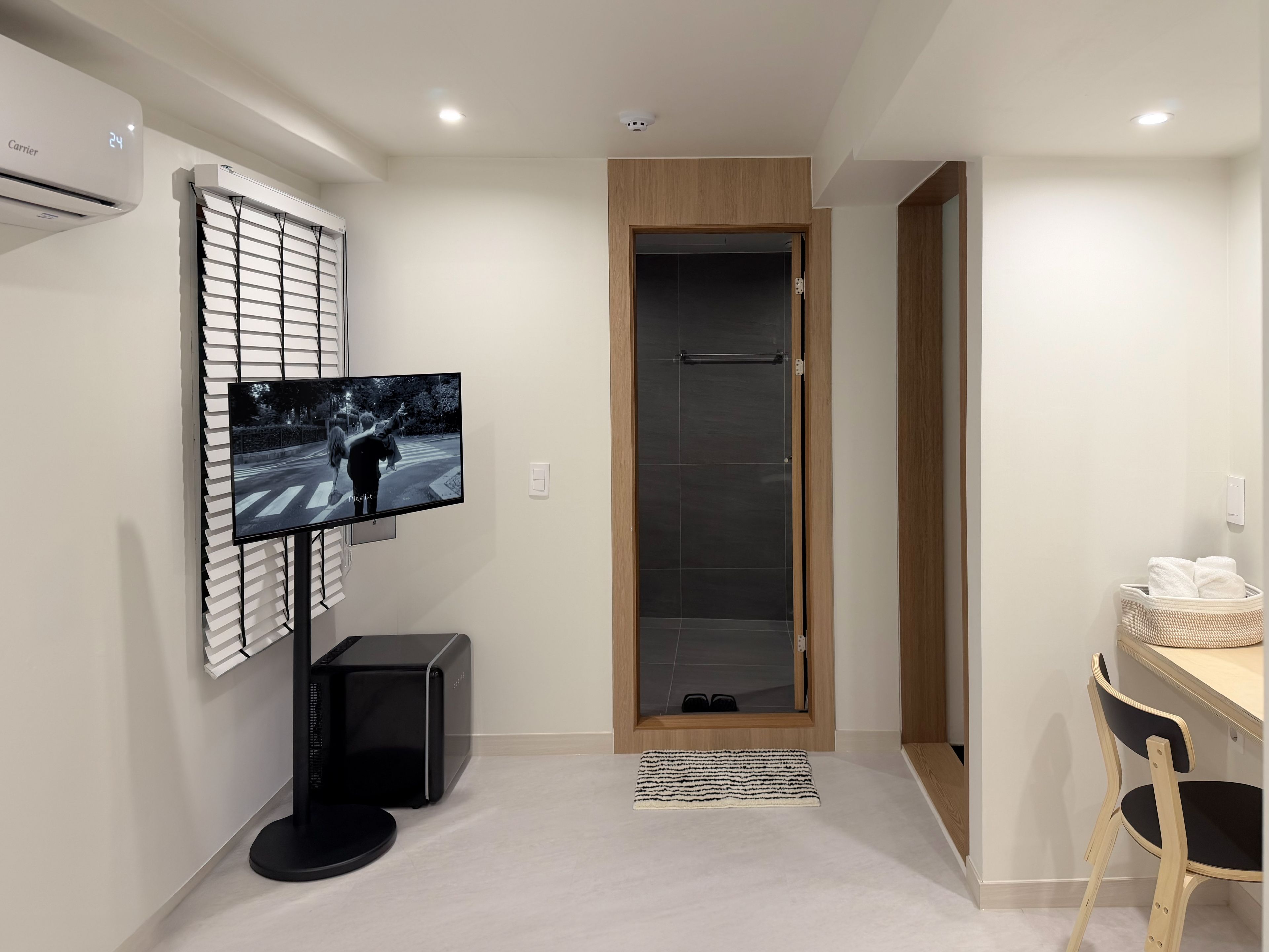 A modern hotel room with a flat-screen TV mounted on a wall, a wooden door, a small table with a chair, and a basket of towels.