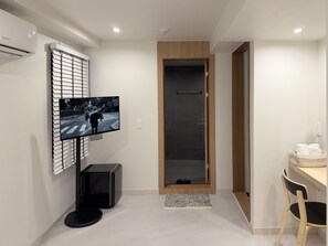 A modern hotel room with a flat-screen TV mounted on a wall, a wooden door, a small table with a chair, and a basket of towels. - Stay Zam Hotel (Busan)