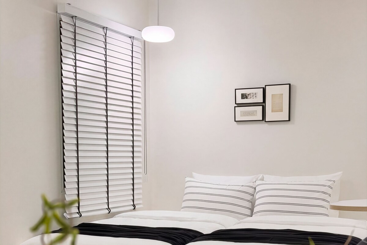 A bedroom with a bed, bedside table, wall-mounted pictures, and a window with blinds.