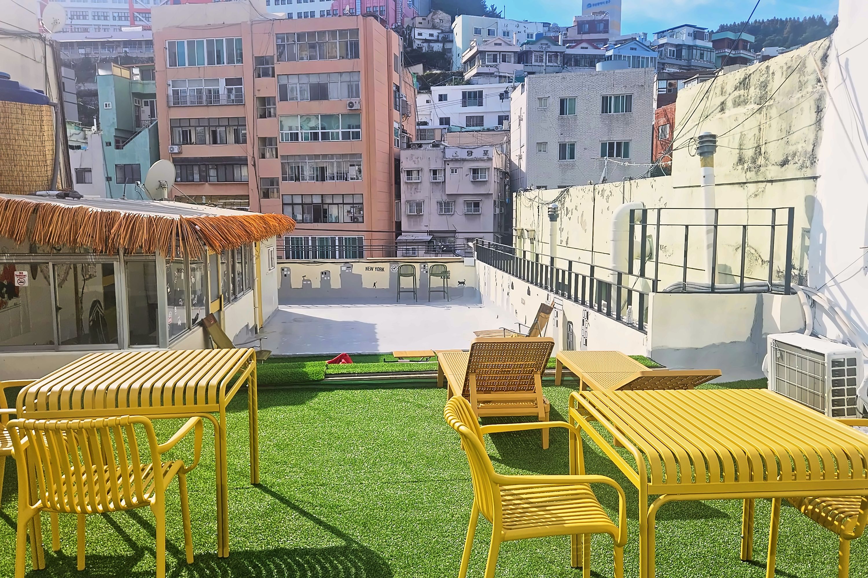 Una terraza en la azotea con sillas y mesas amarillas, un pequeño jardín y vistas de los edificios circundantes.