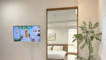 Habitación de hotel con cama, un espejo grande, un televisor mostrando una escena de cafetería y una planta en maceta.