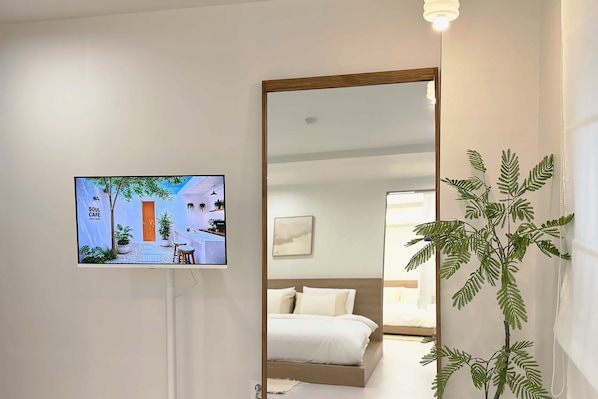 A hotel room with a bed, a large mirror, a television displaying a café scene, and a potted plant.