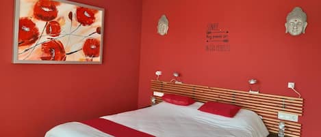 A bedroom with a bed, red walls, a painting, and two Buddha statues.