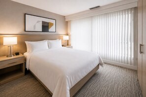 Ein Hotelzimmer mit einem großen Bett, zwei Nachttischen mit Lampen, einem gerahmten Kunstwerk über dem Bett und einem Fenster mit Jalousien.