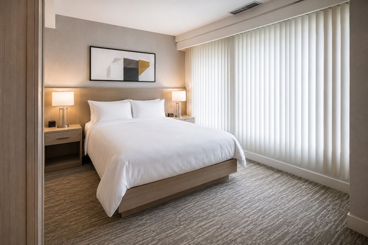 Ein Hotelzimmer mit einem großen Bett, zwei Nachttischen mit Lampen, einem Kopfteil und einem Fenster mit Jalousien.