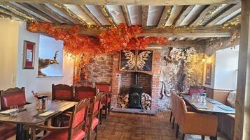 Interior de restaurante con paredes de ladrillo, vigas de madera y una chimenea. Las mesas están dispuestas con sillas tapizadas en rojo.