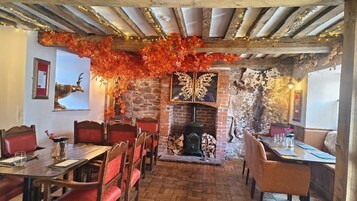 Interior de restaurante con paredes de ladrillo, vigas de madera y una chimenea. Las mesas están dispuestas con sillas tapizadas en rojo.