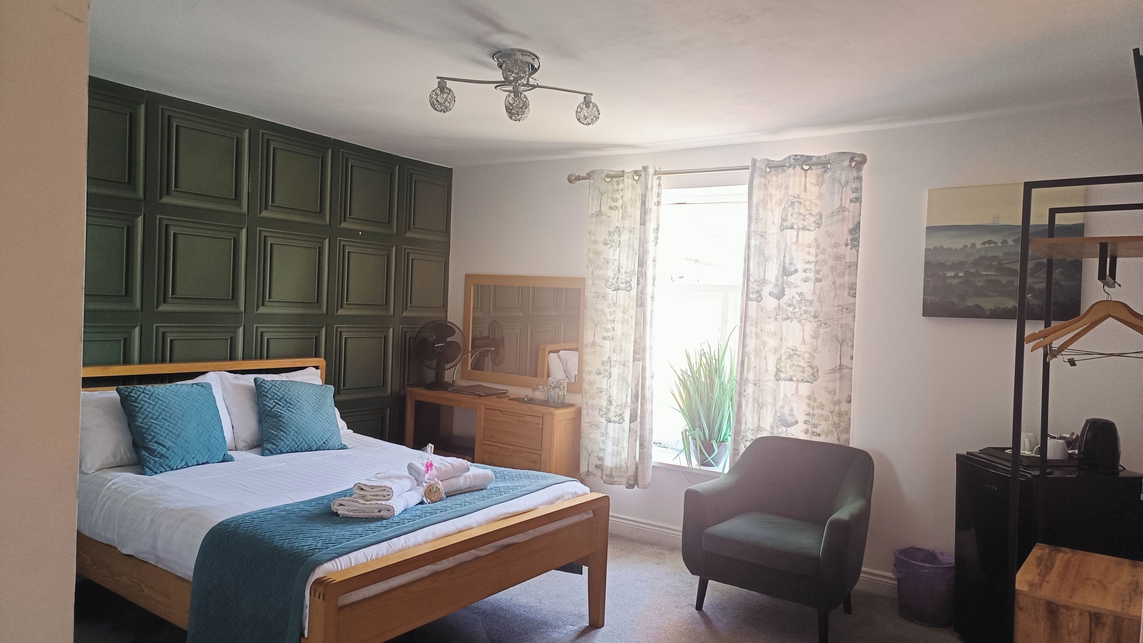 Ein Schlafzimmer mit einem Bett, einem Sessel, einer Kommode, einem Spiegel und einem Fenster mit Vorhängen.