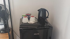 Un refrigerador negro compacto con una tetera y un ventilador encima, ubicado en un rincón de una habitación con un radiador y un armario de madera.