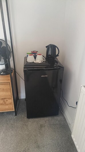 Un refrigerador negro compacto con una tetera y un ventilador encima, ubicado en un rincón de una habitación con un radiador y un armario de madera.