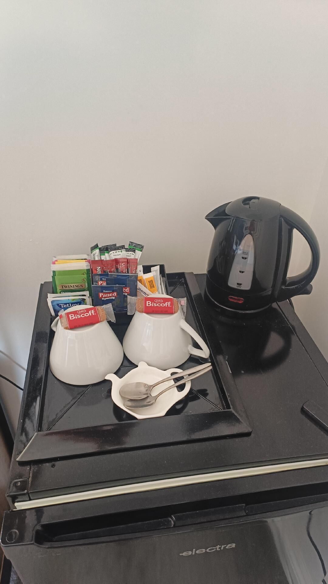 A kitchenette with a coffee maker, two white teapots, a spoon, and various food items including snacks and a Biscoff bar.