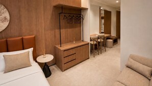 Moderni hotellihuone, jossa on sänky, pieni pyöreä pöytä, puinen kaappi, oleskelualue pöydällä ja tuoleilla sekä naulakko.