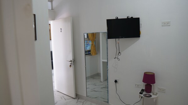 A room with a door leading to another space, a glass shower enclosure, a mounted television, and a small table with a lamp and remote control.