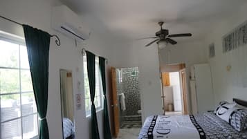 Un dormitori amb un llit, un ventilador de sostre, una finestra amb cortines i una porta que dona a un bany.