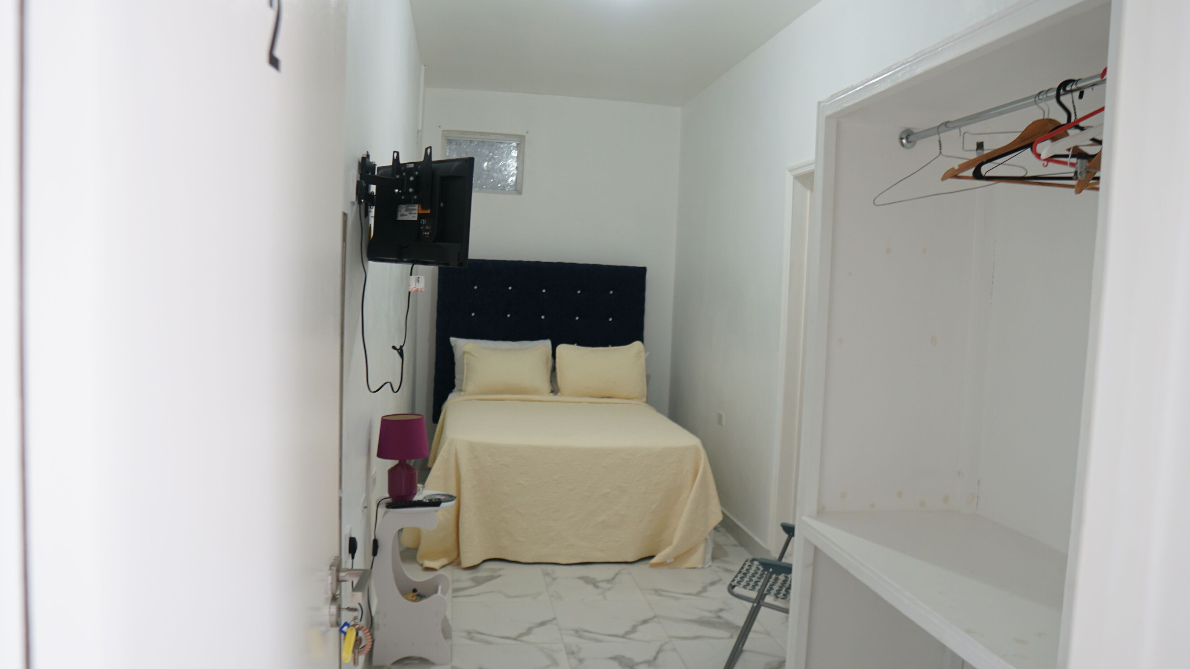 Una camera d'albergo piccola e stretta, con un letto, un comodino, una lampada, uno specchio e una TV a muro.