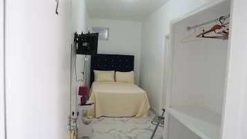 Quarto de hotel pequeno e estreito, com uma cama, mesa de cabeceira, luminária, espelho e televisão fixada na parede.