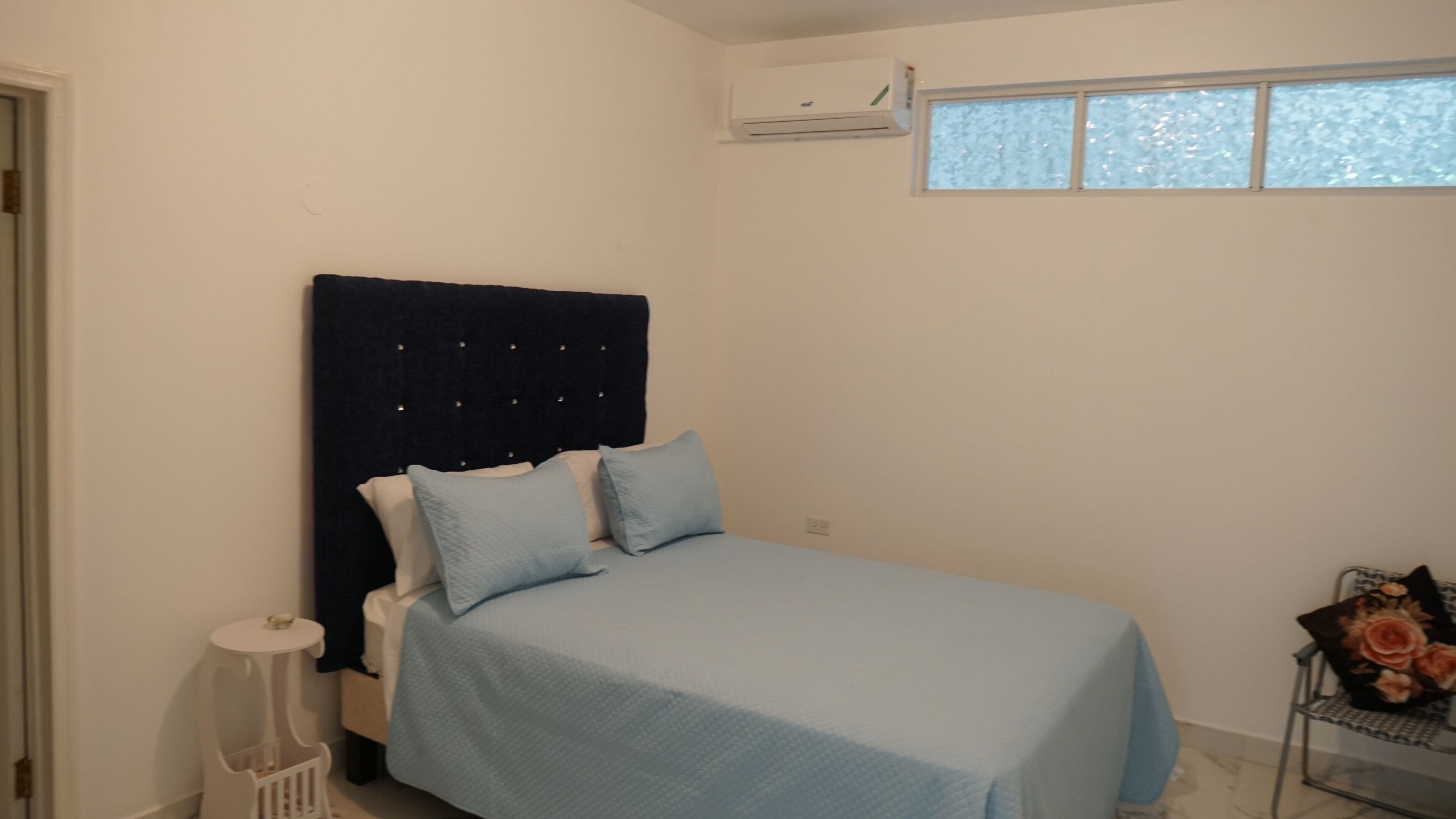 A bedroom with a bed, a window, an air conditioning unit, and a chair with a floral cushion.