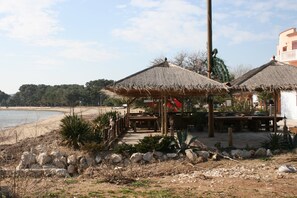 Ein Restaurant direkt am Strand mit reetgedeckten Sonnenschirmen und Holztischen, mit Blick auf den Sandstrand und das Meer.