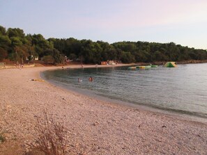 A pebbled beach with people swimming and a forested area in the background. - Schöne Ferienwohnung "Scampi" in ruhigem Stadtteil von Pula mit 2 Schlafzimmer (Pula)