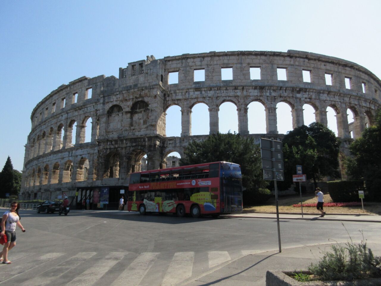 Un amphithéâtre historique, doté de nombreuses arcades, avec un autobus à deux étages rouge en premier plan.