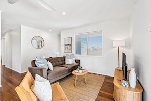 A modern living room with a brown sofa, a round mirror, a framed picture, a window with blinds, and a small round table with a lamp.
