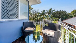Un balcón con mobiliario de mimbre y una mesa de vidrio, con vista a un jardín con palmeras.