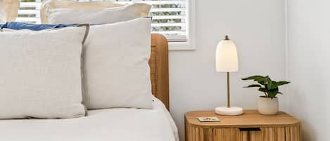 A neatly made bed with pillows and a blue blanket, a bedside lamp, a small wooden nightstand with a plant, and a framed picture on the wall.