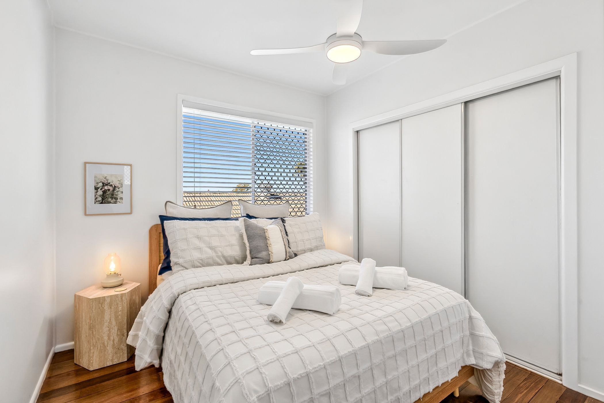 A bedroom with a bed, a bedside table with a lamp, a window with blinds, and a ceiling fan.