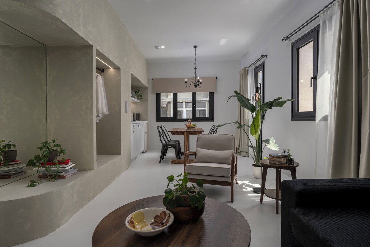 A modern living room with a dining area, a kitchen in the background, and a large window with curtains.