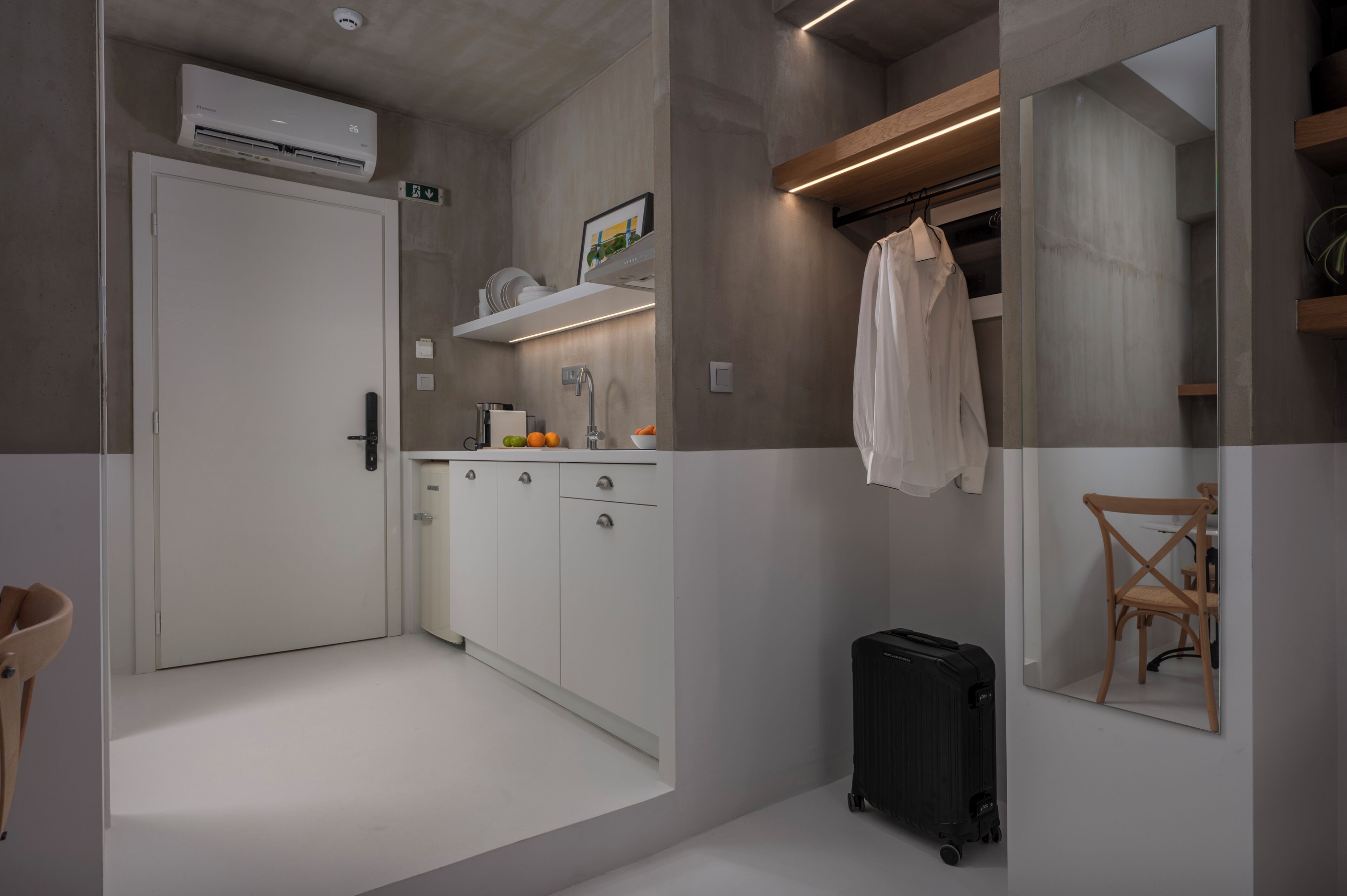Una habitación de hotel moderna con kitchenette, un espejo, una maleta y una silla de madera.
