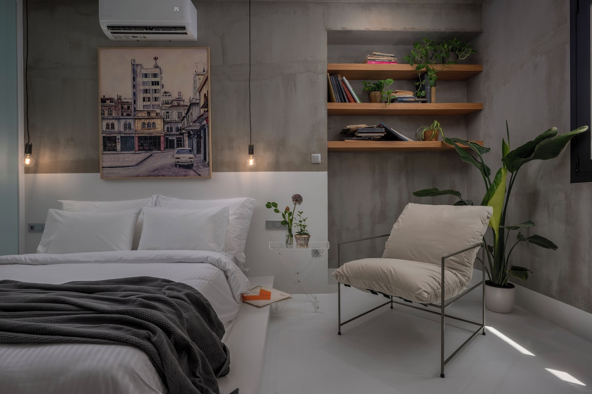 A bedroom with a bed, a chair, a shelf with books and plants, a painting on the wall, and an air conditioning unit.