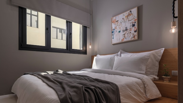 A modern bedroom with a large window, a bed with white linens, a gray throw blanket, a wooden bedside table with a plant, and a wall-mounted abstract painting. - Binome Suites (Thessaloniki)