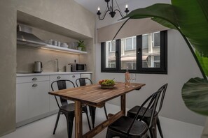 A compact kitchen with white cabinets, a wooden dining table, and a window with black shutters. - Binome Suites (Thessaloniki)