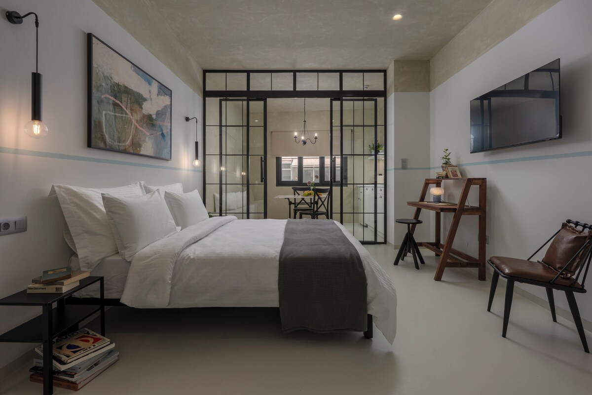 A bedroom with a bed, bedside table, and a chair. There is a desk with a chair and a TV mounted on the wall.