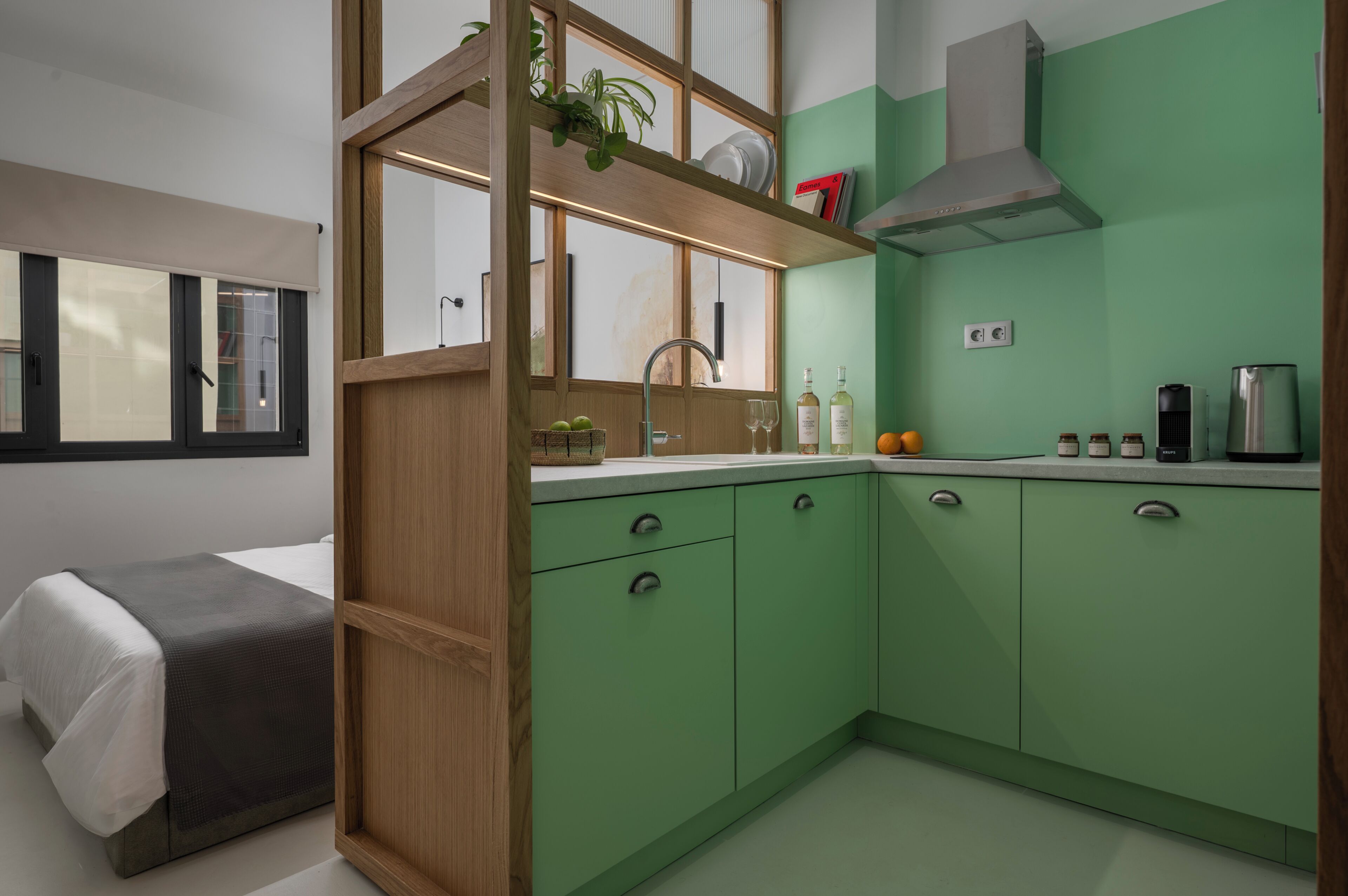 Une cuisine compacte avec des meubles de couleur verte, une cloison en bois et une fenêtre avec des stores.