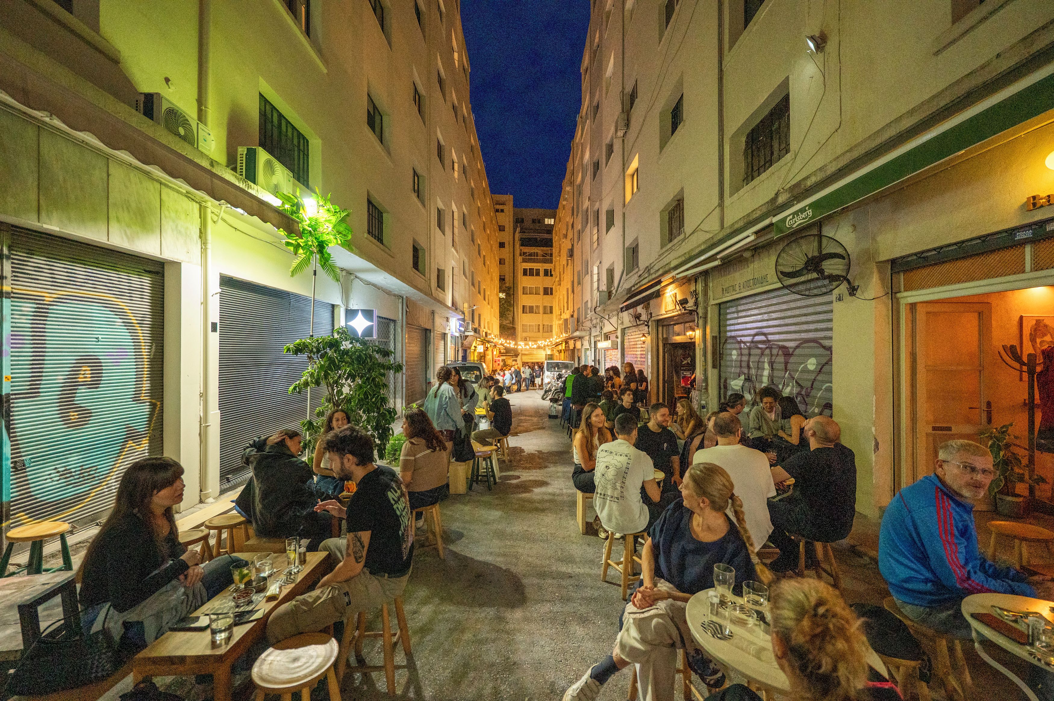 Ein Straßencafé mit Tischen und Stühlen, umgeben von Gebäuden mit heruntergelassenen Rollläden.