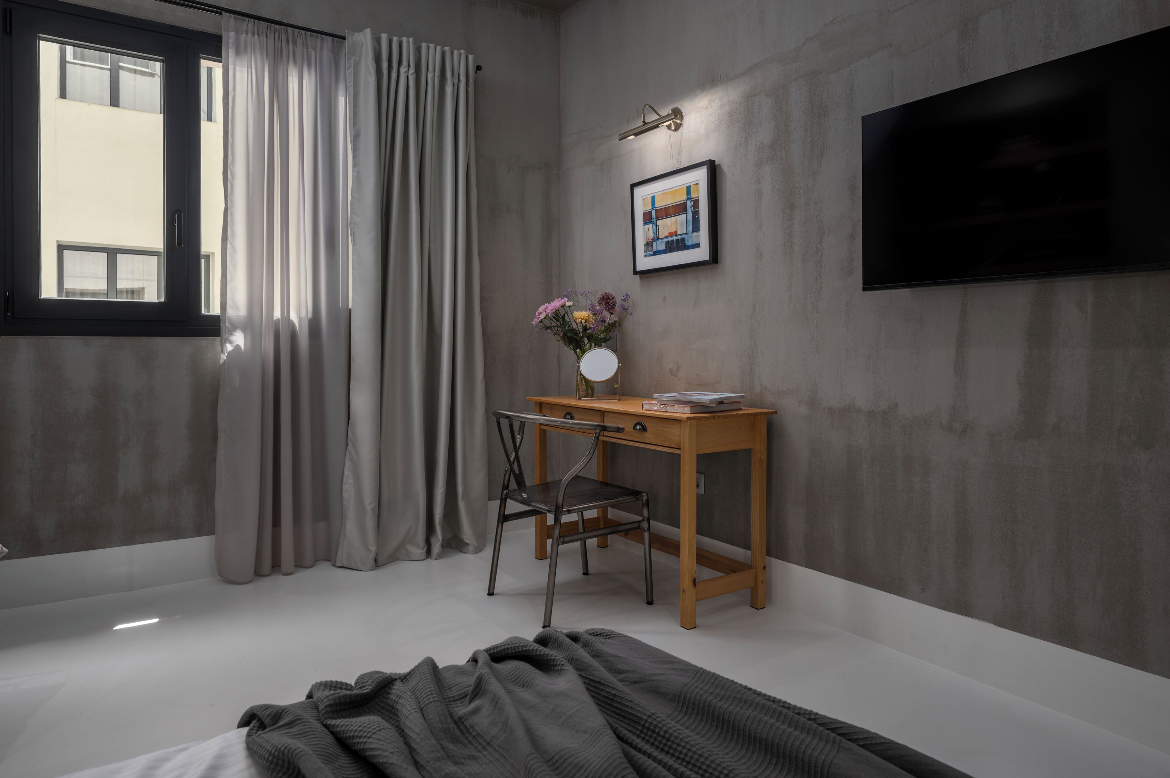 A bedroom with a bed, a desk with a chair, a TV on the wall, and a window with curtains.