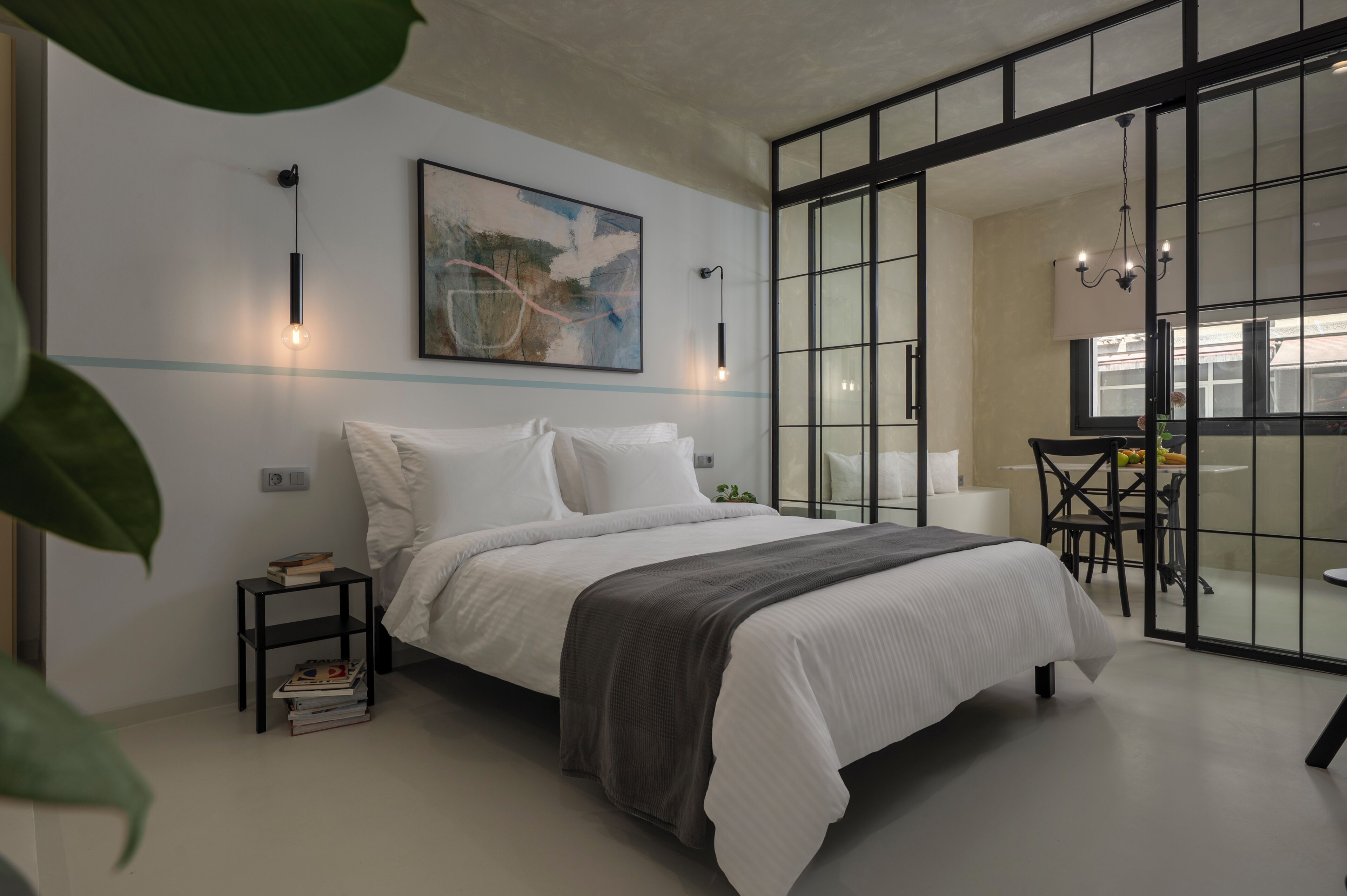 A modern bedroom with a large bed, a side table, a painting, and a sliding glass door leading to a dining area.