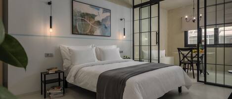 A modern bedroom with a large bed, a side table, a painting, and a sliding glass door leading to a dining area.