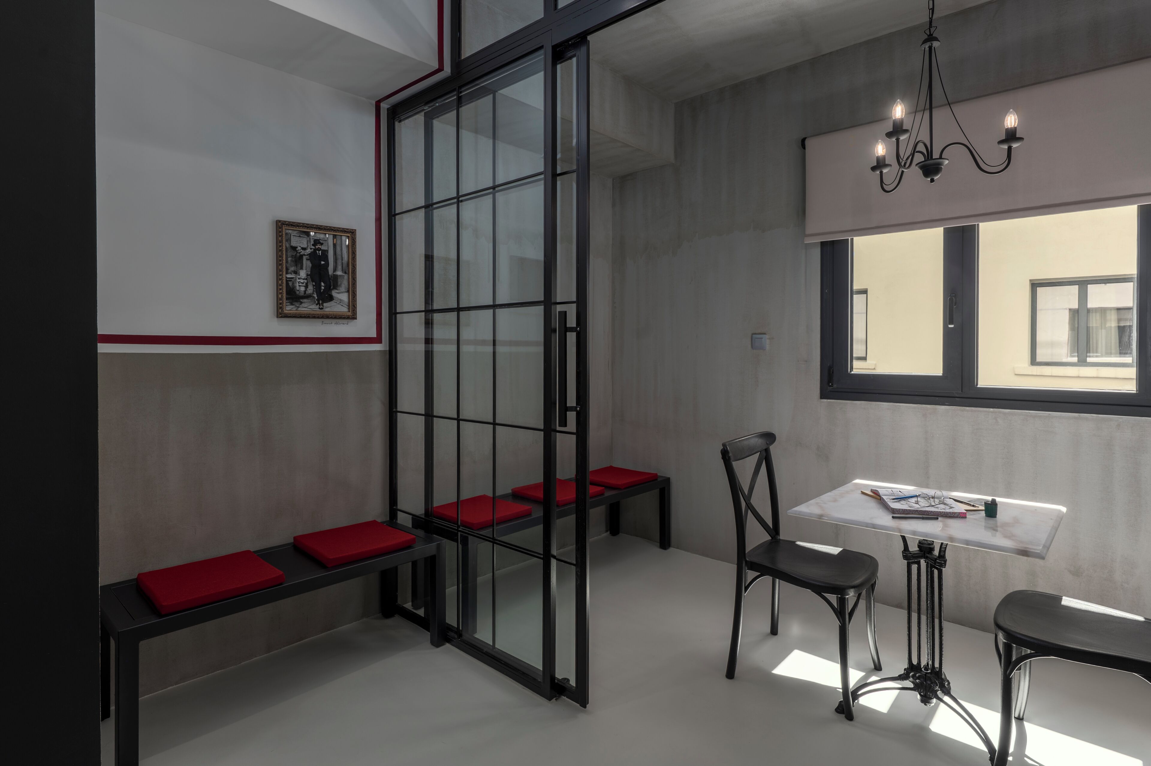A modern interior with a glass partition, red cushioned seating, a table with chairs, and a chandelier.