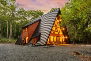 Eine moderne, dreieckige Holzhütte mit geneigtem Dach, großen Fenstern und einer Schotterauffahrt.