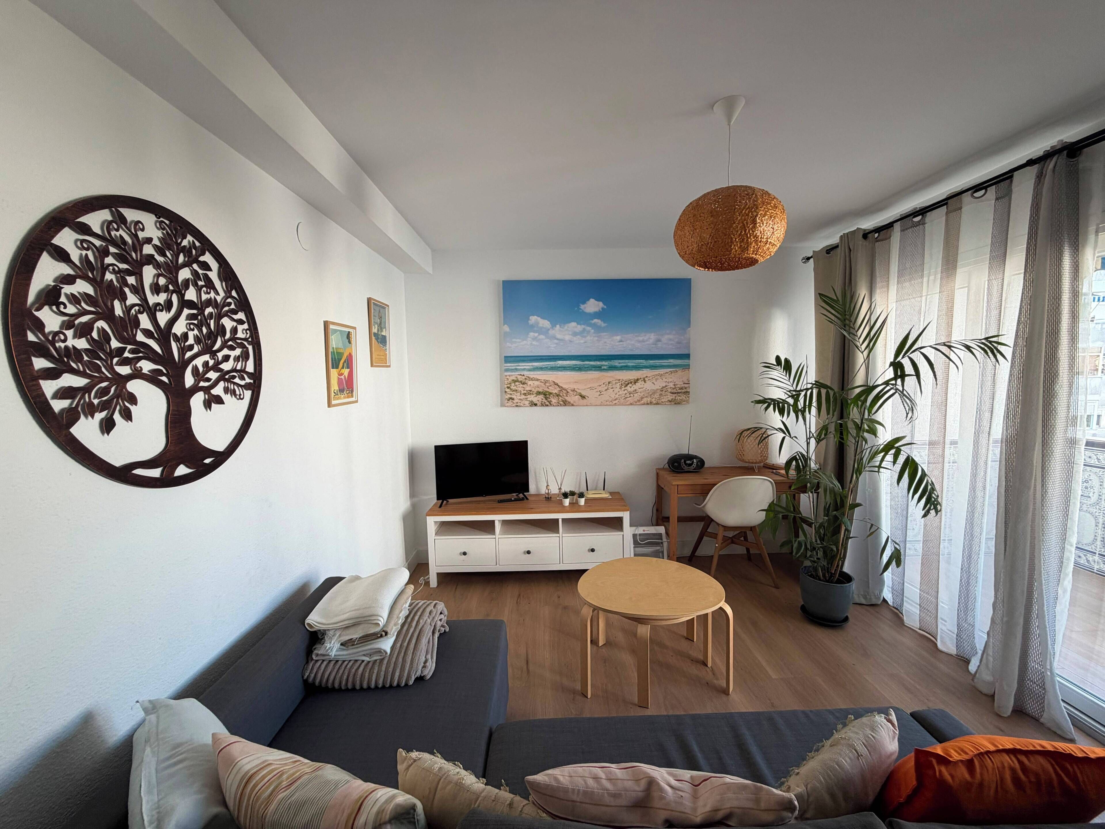 Ein Wohnzimmer mit einer Couch, einem Couchtisch, einem Fernseher, einem Strandbild, einer Baumdekoration und einer Pflanze.