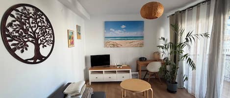 Ein Wohnzimmer mit einer Couch, einem Couchtisch, einem Fernseher, einem Strandbild, einer Baumdekoration und einer Pflanze.