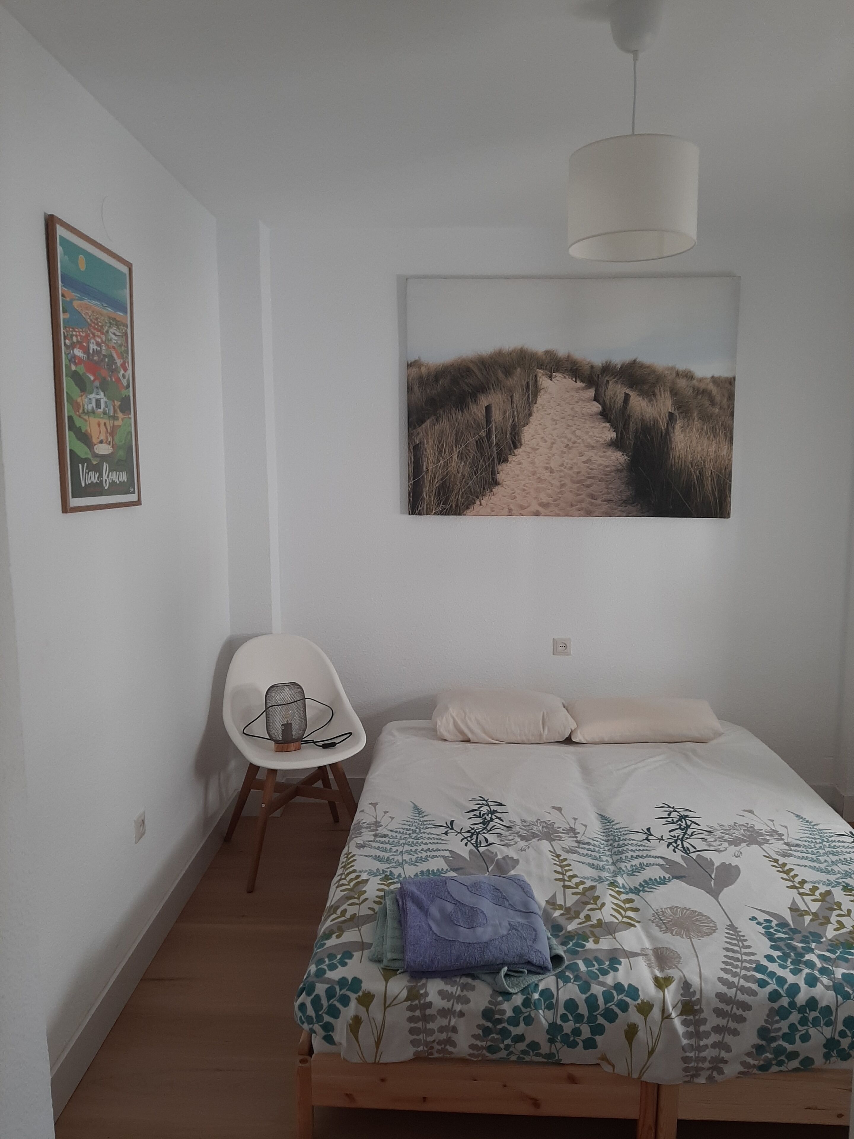 A bedroom with a bed, a chair, a picture on the wall, and a hanging light fixture.