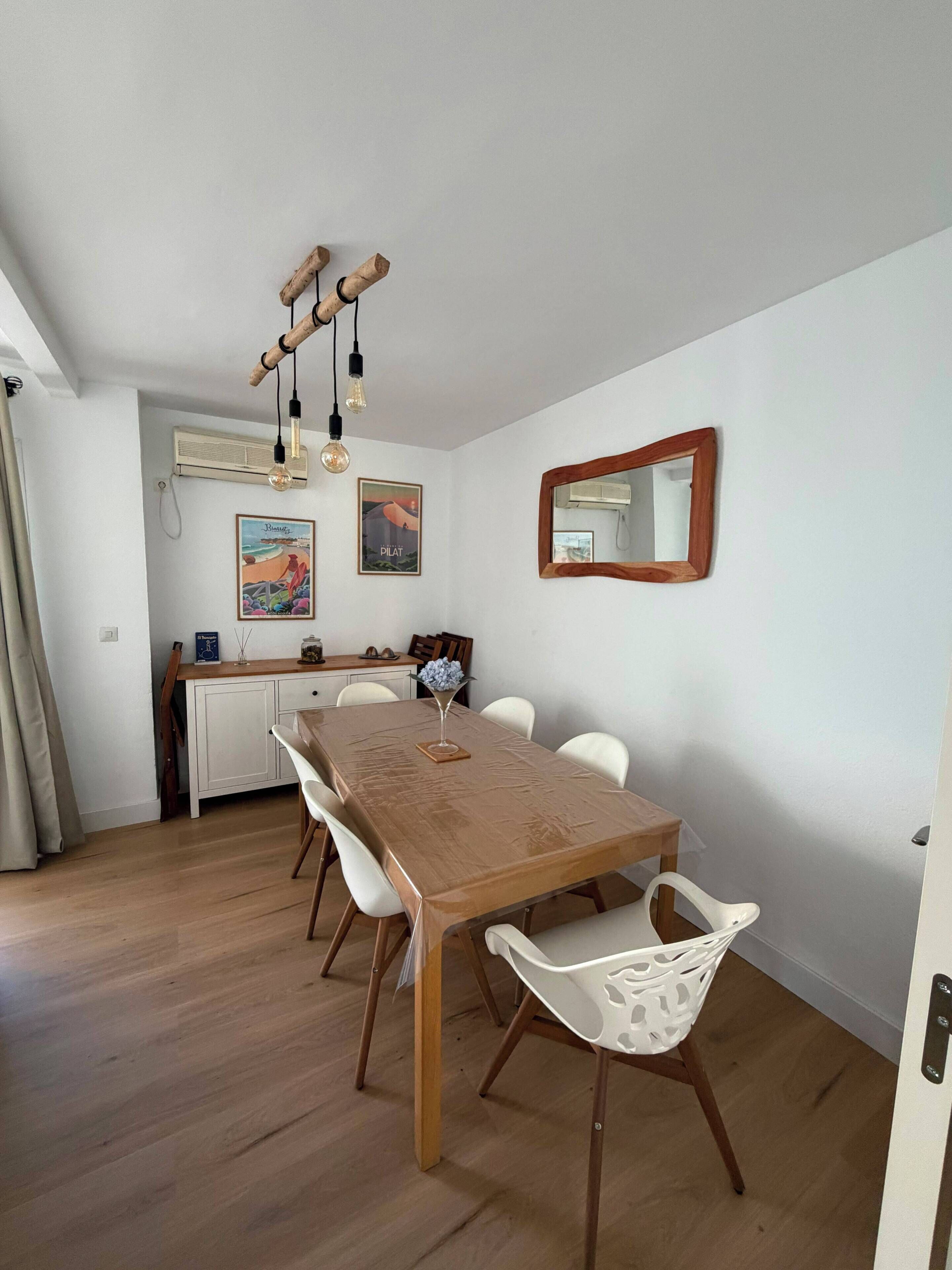 A dining area with a wooden table and white chairs, a hanging light fixture, and wall-mounted air conditioning units.