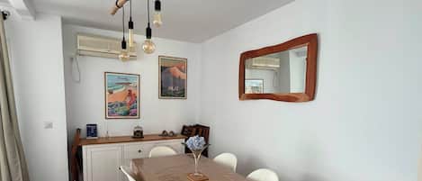 A dining area with a wooden table and white chairs, a hanging light fixture, and wall-mounted air conditioning units.