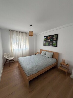 Ein Schlafzimmer mit einem hölzernen Bett, einem Sessel, einem kleinen Tisch, einem an der Wand befestigten Bilderrahmen und einer Pendelleuchte.