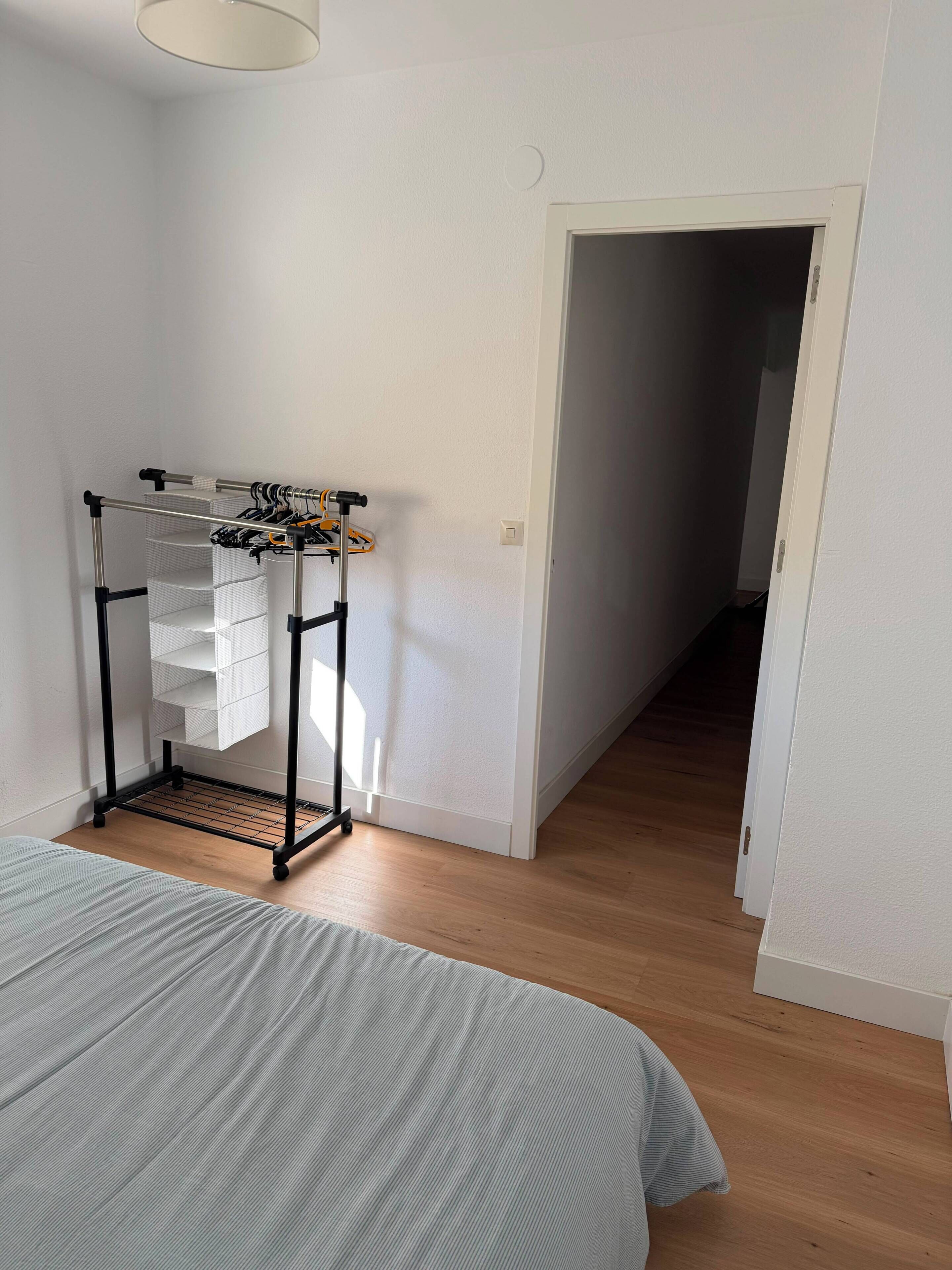 Une chambre avec un lit, un portant en métal où sont suspendus des vêtements, et un couloir menant à une autre pièce.